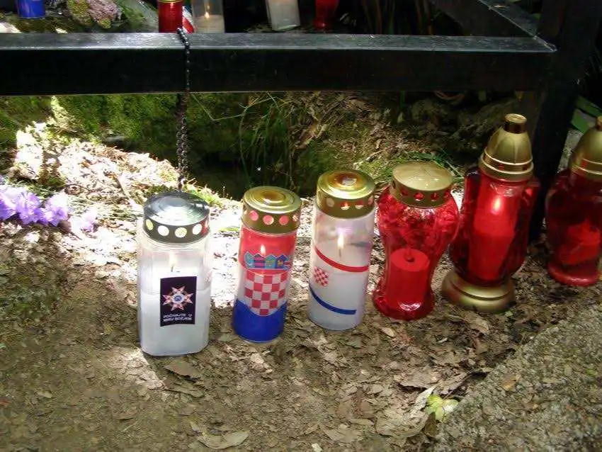 Upaljeni lampioni s hrvatskim obilježjima postavljeni uz metalnu ogradu na šumskom tlu, u znak sjećanja na žrtve.