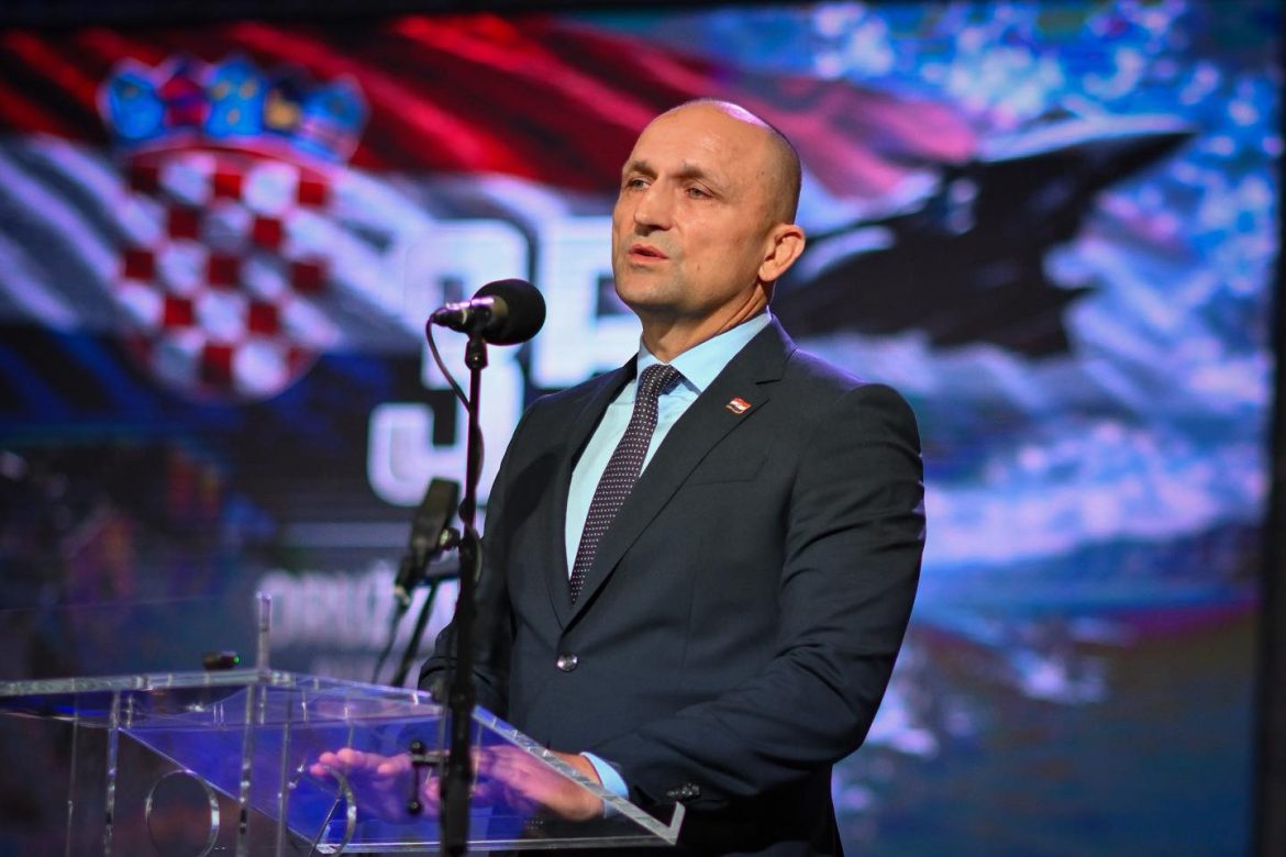 Man in a dark suit delivering a speech at a clear lectern on a stage, with a patriotic backdrop and flag pin on his lapel.
