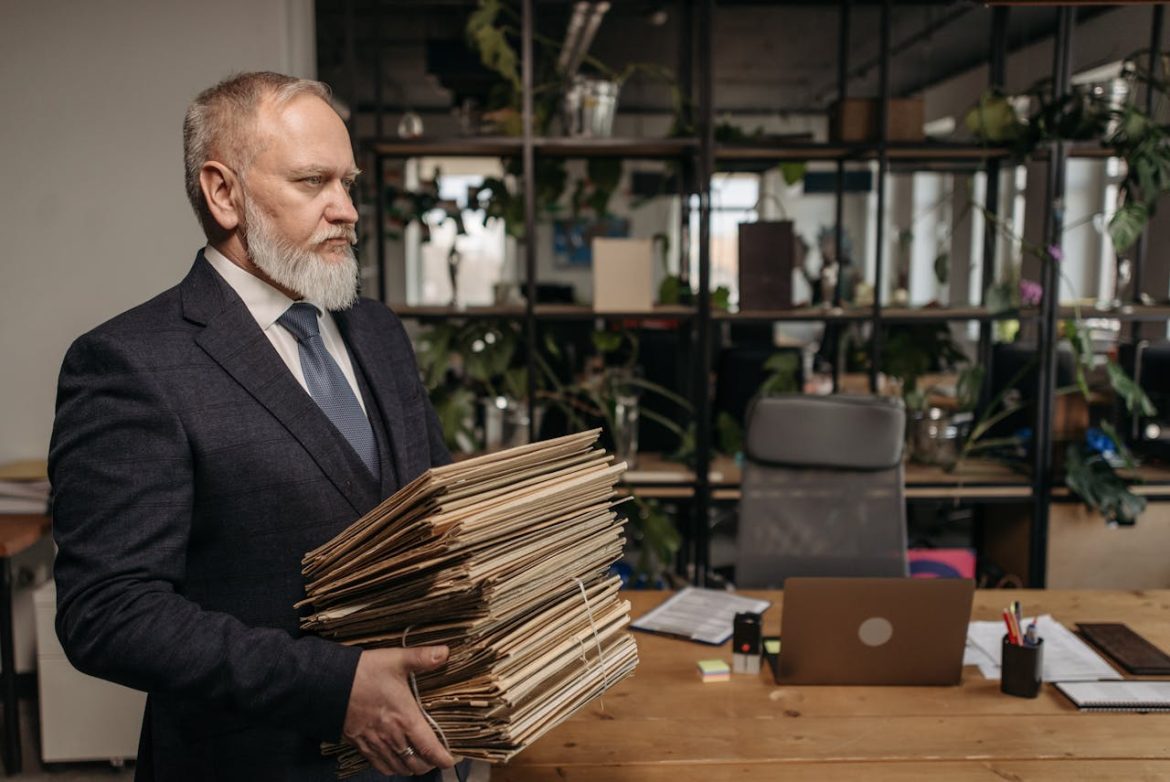 Businessman in a dark suit holding a large stack of manila folders in a modern office.