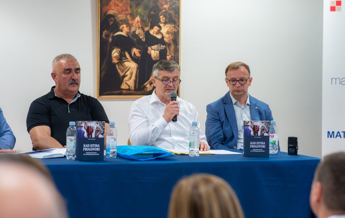 Three men seated at a panel table during a press conference; the center man speaks into a microphone, flanked by two colleagues, with water bottles and books in front and a painting on the wall behind.