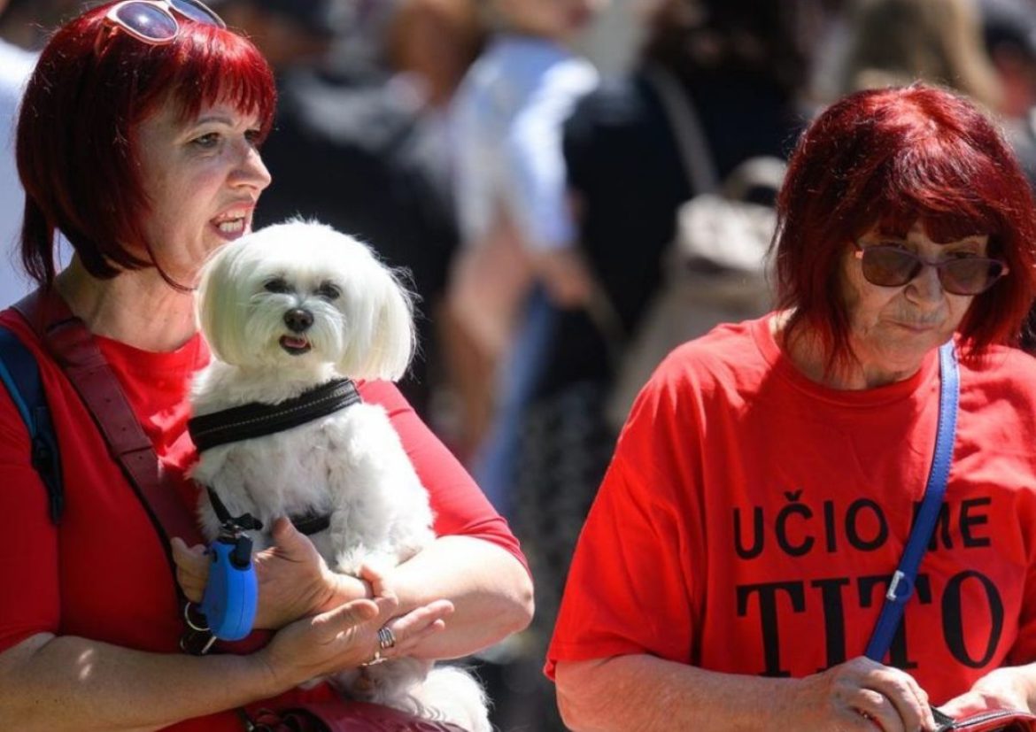 Two women in red shirts outdoors, one holding a small white fluffy dog with a harness.