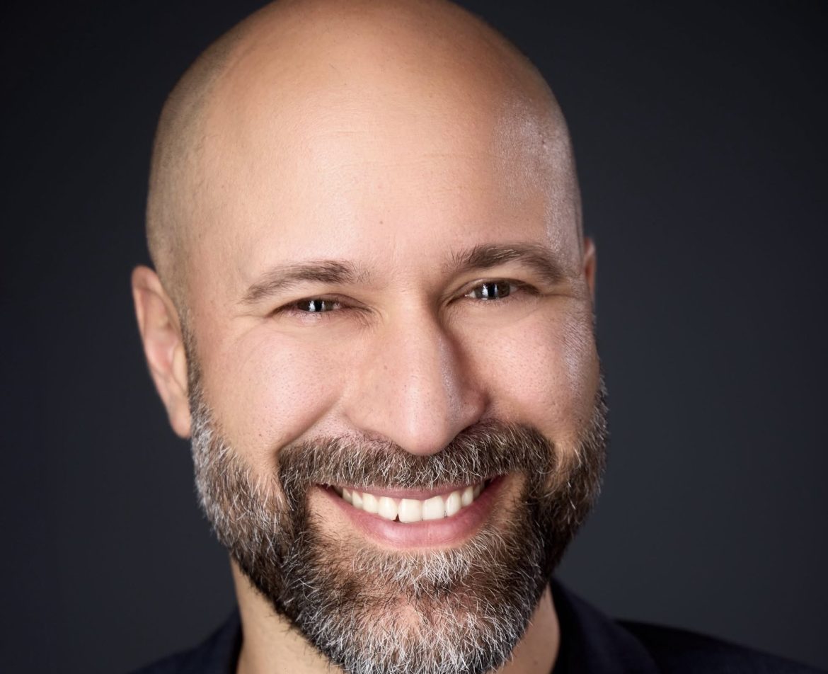 Close-up portrait of a smiling man with a gray beard and shaved head against a dark background, showing his teeth.