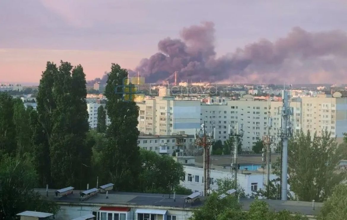 City skyline with tall buildings and a thick dark smoke plume rising behind them at dusk, viewed over trees and rooftops in the foreground.