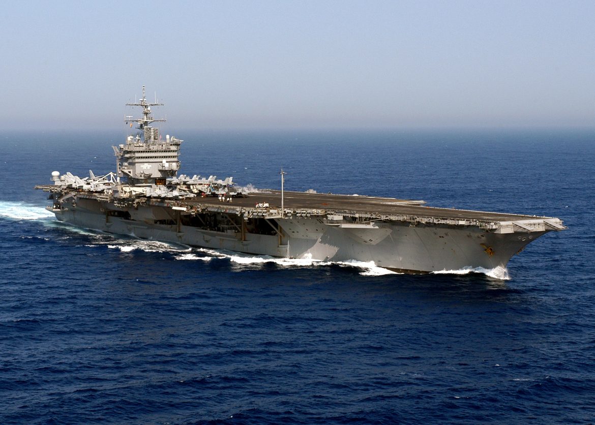 Flight deck of a large aircraft carrier at sea with airplanes parked along the edge and a control tower rising above the deck.