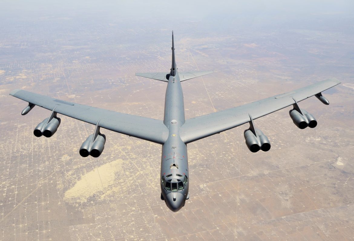 Top-down view of a large gray bomber (B-52) with eight engines, flying over a dry desert landscape.