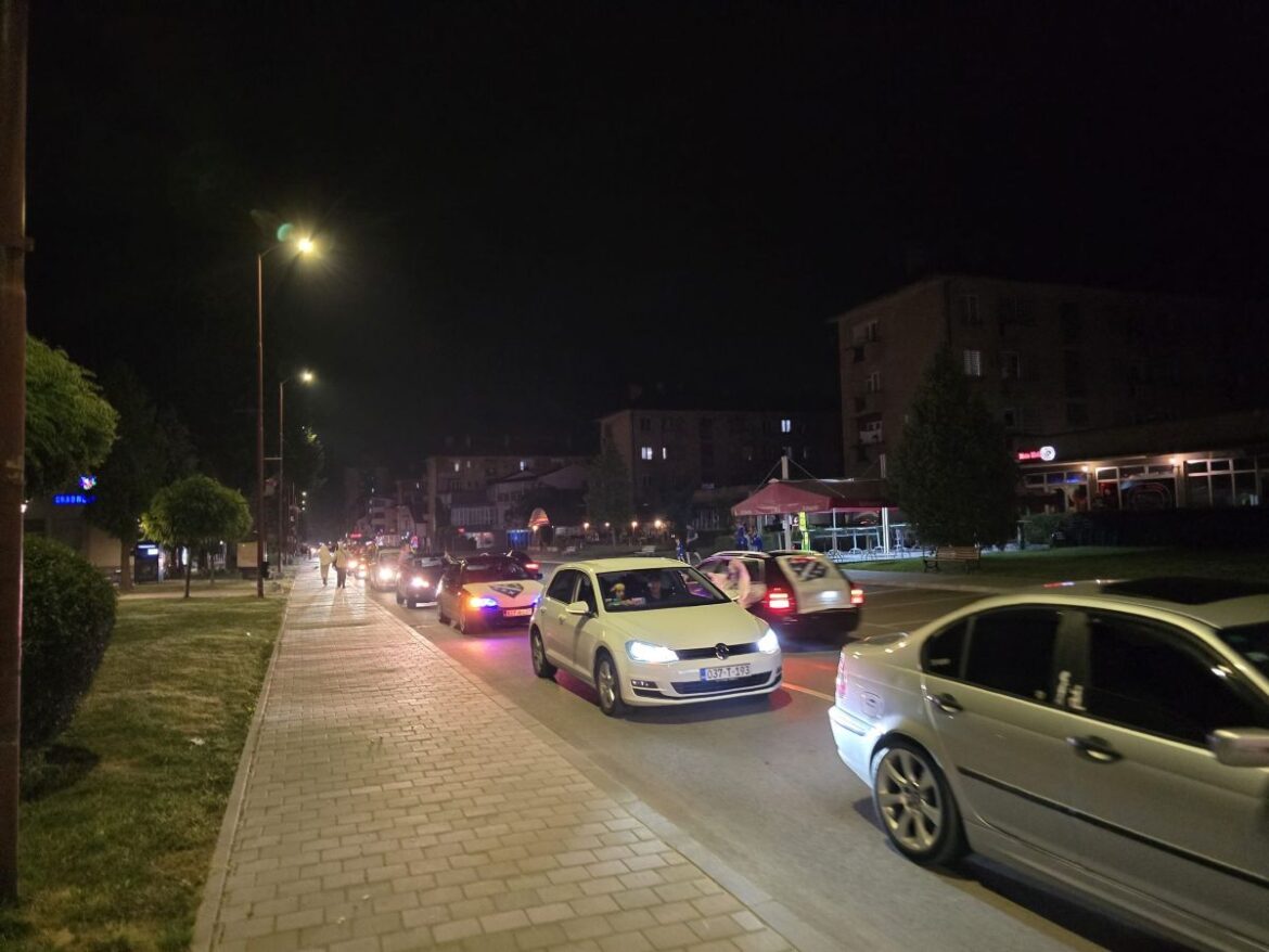 Night street scene at night with cars on a two-lane road, a brick sidewalk on the left, and buildings in the background under streetlights.