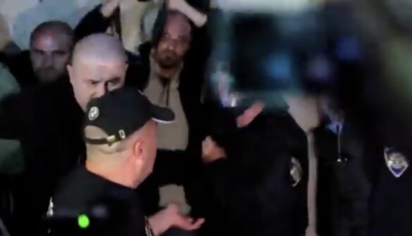 Police officers surround a man in a crowded indoor scene, one officer reaching toward him as others watch.