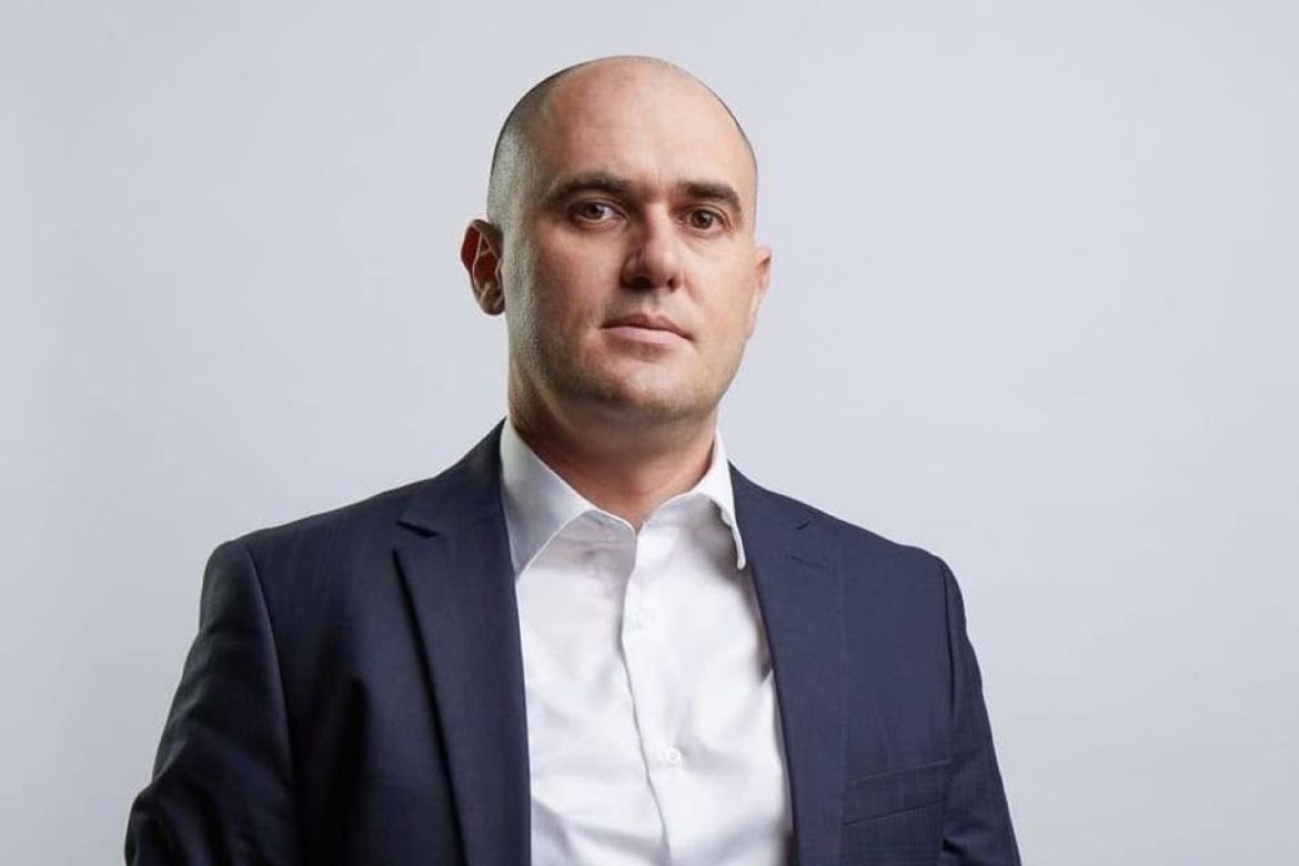 Professional portrait of a bald man in a dark suit and white shirt, looking at the camera against a light gray background.