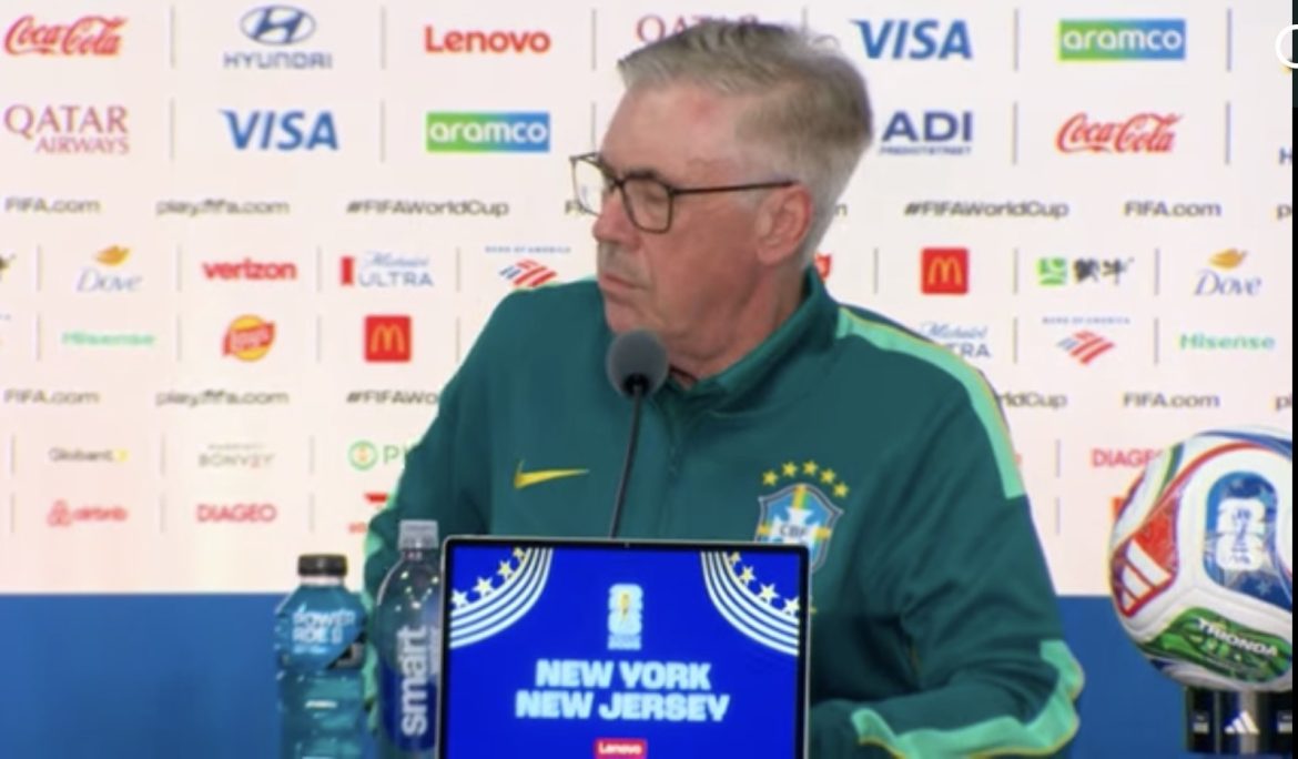 Brazil national team coach at a press conference, wearing a green tracksuit, with a laptop showing 'New York New Jersey' and a soccer ball nearby