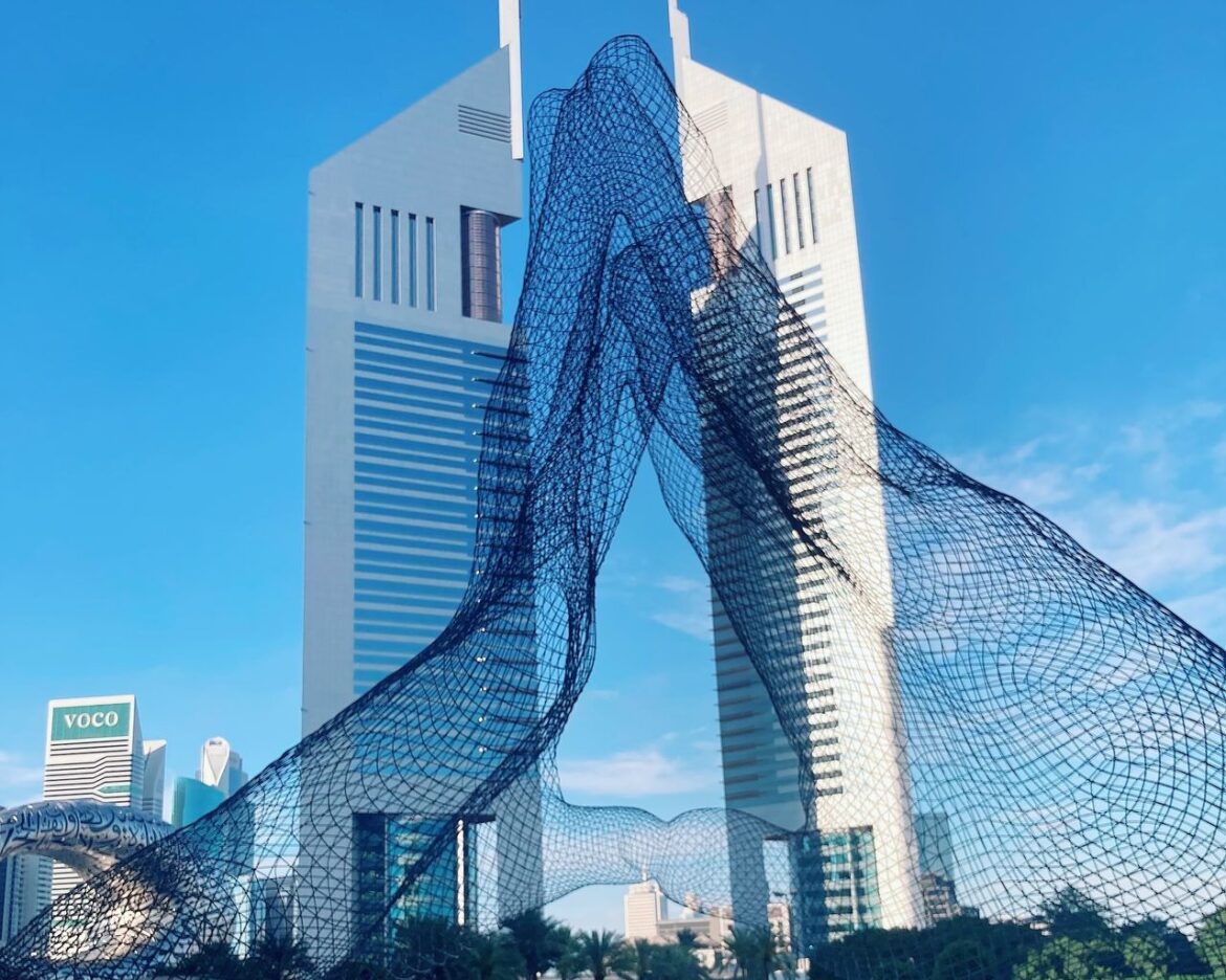 Blue wireframe sculpture spiraling between modern skyscrapers in a city plaza against a clear blue sky.