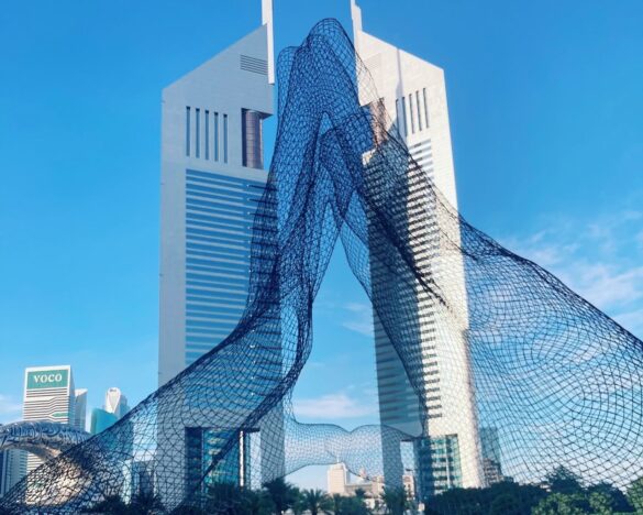 Blue wireframe sculpture spiraling between modern skyscrapers in a city plaza against a clear blue sky.