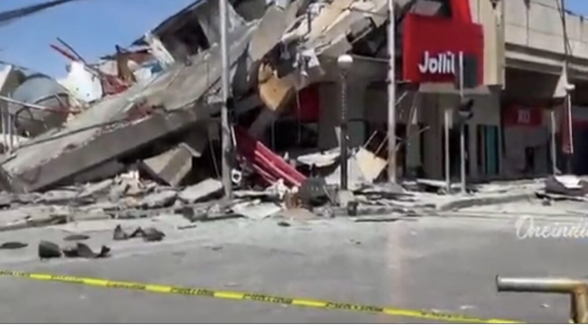 Street scene showing a collapsed building with rubble and debris across the sidewalk and road, with yellow caution tape in the foreground and a red storefront sign visible.