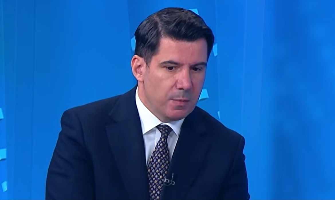 A man in a dark suit and patterned tie sits against a bright blue studio backdrop, speaking during a TV interview.