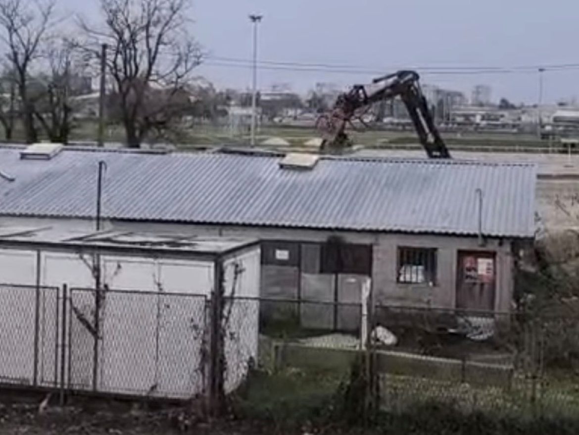 Industrial site with a metal-roofed building, chain-link fence, and a large excavator arm reaching over the roof in the background.