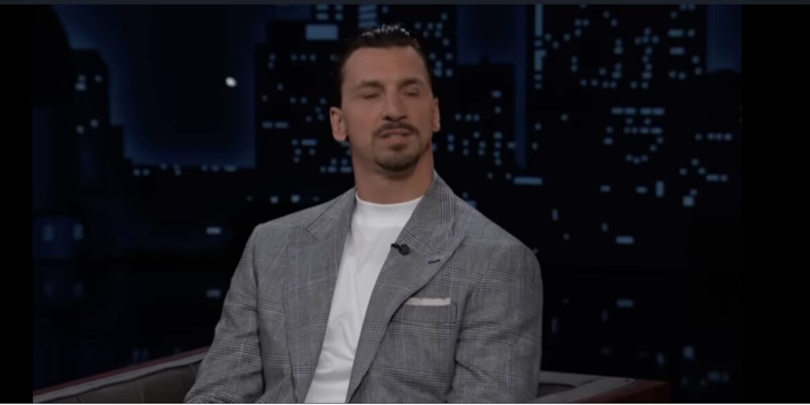 Man in a gray plaid blazer and white shirt sits on a talk-show set with a dark cityscape backdrop and a clip-on microphone visible on his lapel area.