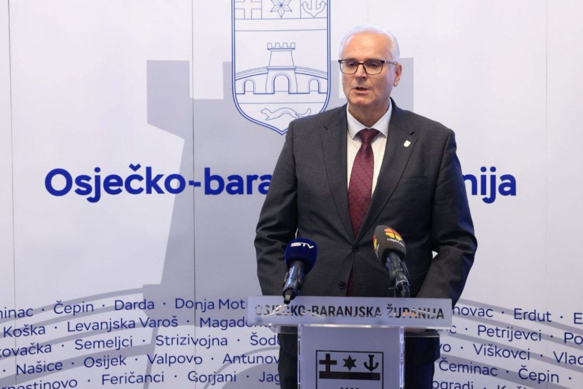 Male official in dark suit and red tie speaks at a podium with microphones, in front of a backdrop reading 'Osječko-baranjska županija' and a city crest.