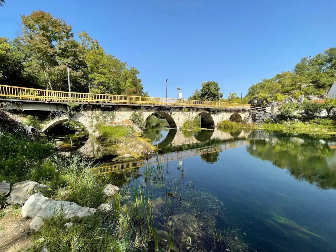 Stone arch bridge with yellow railing spans a calm river, reflected in the water, with trees and a clear blue sky everywhere around.