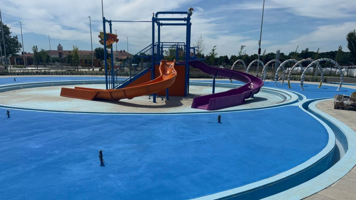 Water playground under construction with blue circular splash pad and orange and purple slides in the center area under a clear sky
