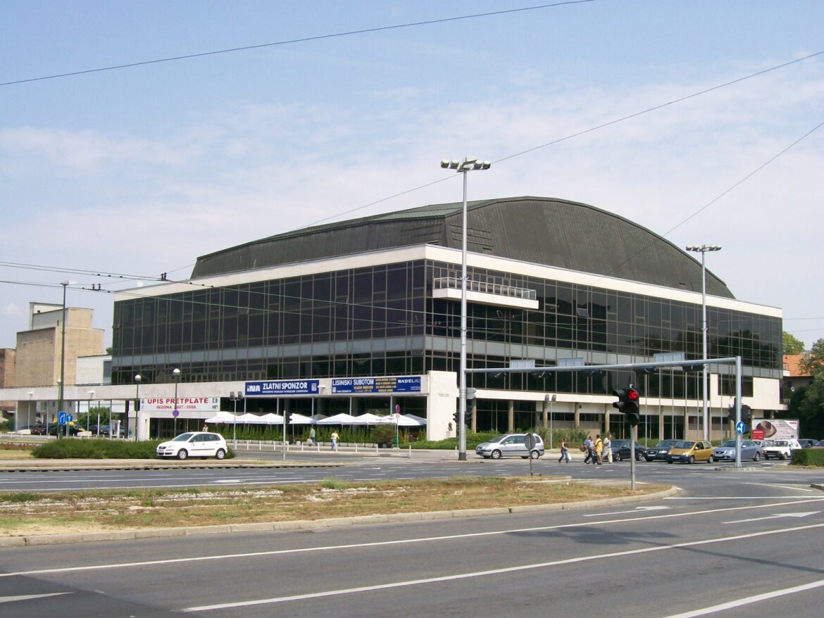 Modern glass-front commercial building with a curved dark roof, seen along a busy road with pedestrians and cars nearby.