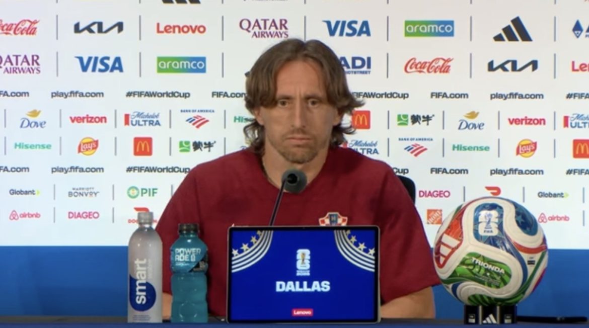 Male football coach at a press conference, laptop displays 'DALLAS' with official branding, a colorful soccer ball on a stand to his right, and water bottles on the table.