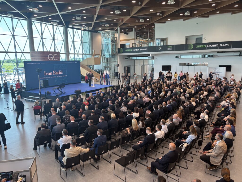 Large indoor conference hall with a blue stage, audience seated in rows, and a speaker podium at center front under a banner reading 'Ivan Radić'.