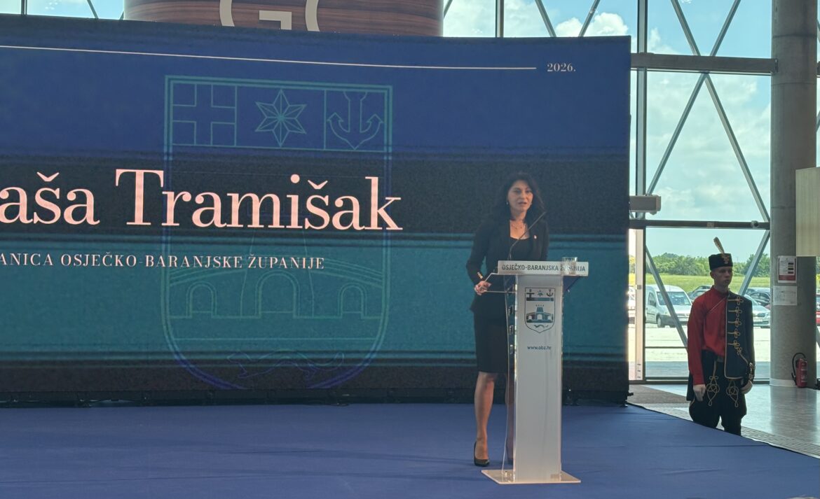 Woman in a black suit speaks at a podium on a blue stage backdrop, with official insignia and 'Osječko-baranjska županija' text visible, ceremonial guard nearby.