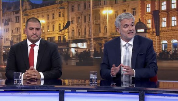 Two men in suits sit at a news desk on a city backdrop, engaged in conversation with microphones attached to their jackets.