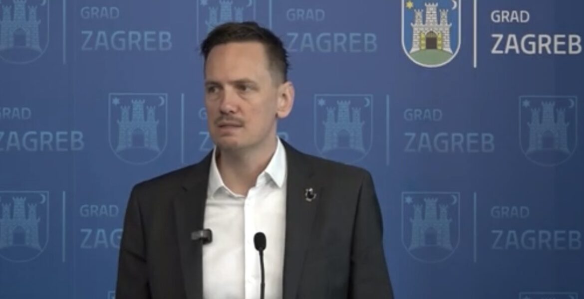 Man in a dark suit speaks at a press conference, two microphones in front of him, backdrop reads 'GRAD ZAGREB' with city crests.