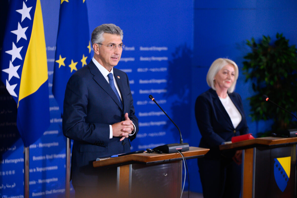 Two officials at a press conference: a man in a dark suit speaking at a podium with microphones, a woman seated at another podium in the background against blue banners and flags.
