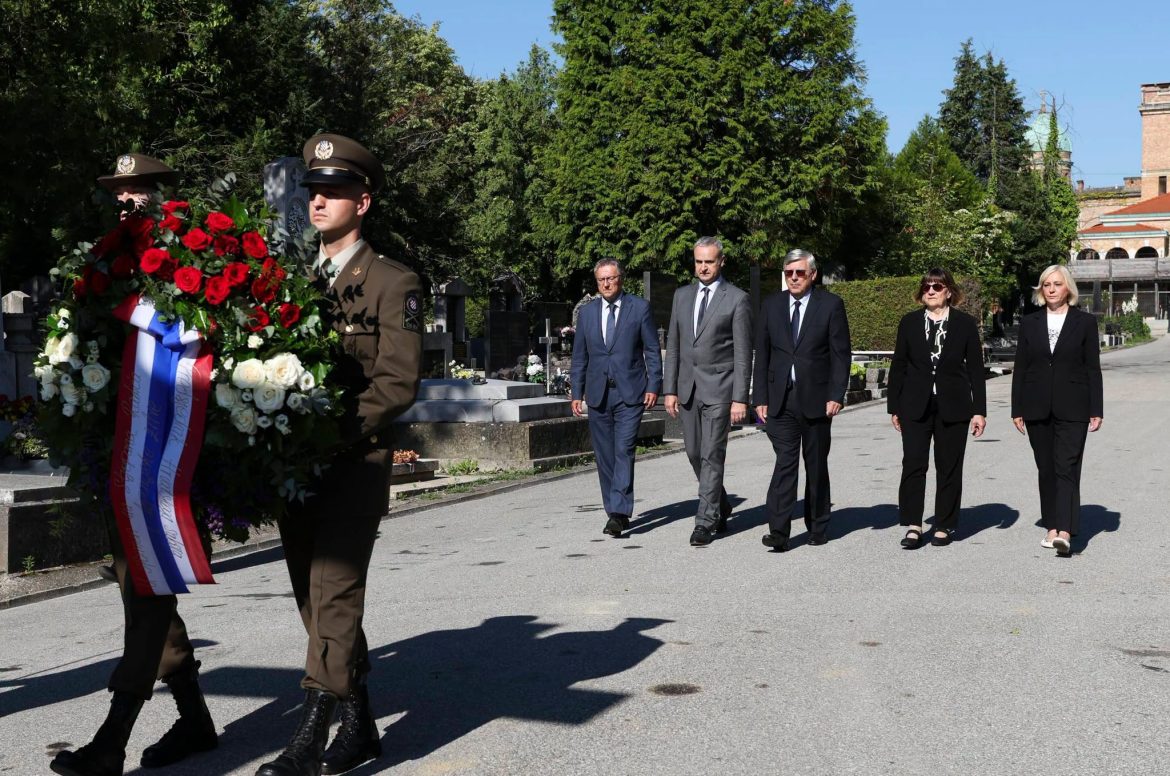 Uniformed soldier carries a large wreath of red and white flowers as five formally dressed civilians walk behind at a cemetery.