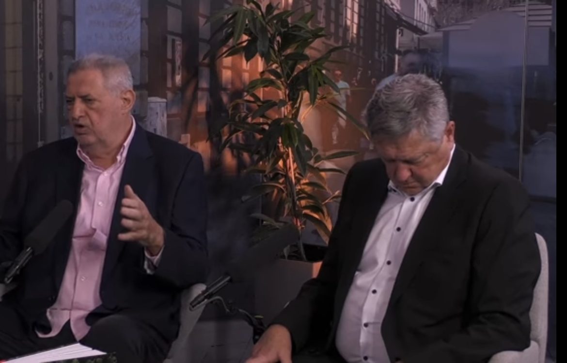 Two men in business suits sit on a talk-show set with microphones in front, one speaking with hand gestures, the other looking down beside a plant backdrop