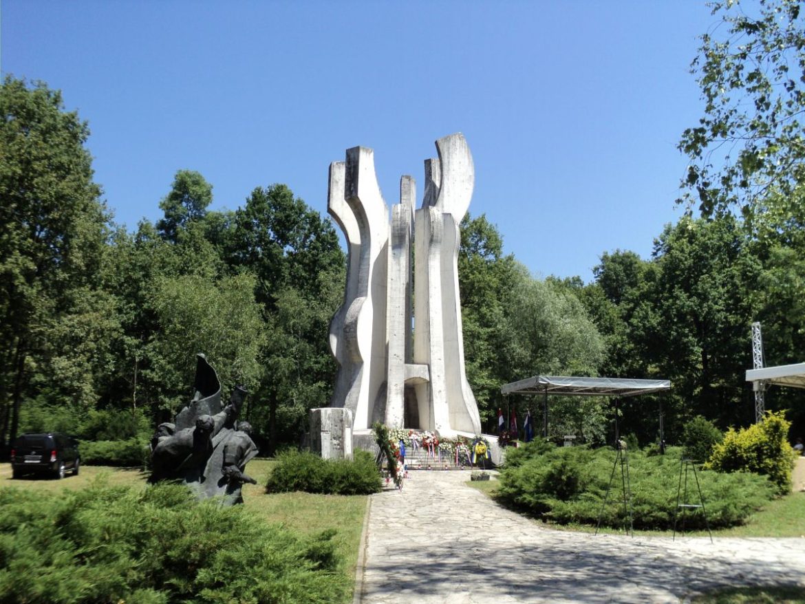 White abstract monument with curved pillars in a park, with statues at left and wreaths at its base
