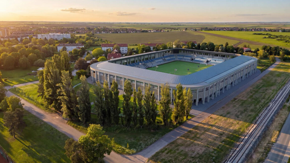 Aerial view of a modern stadium with a green playing field, surrounded by trees and a railway line on the right at sunset.
