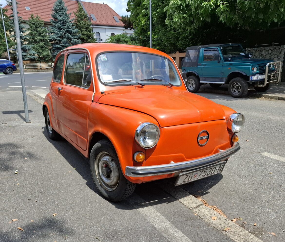 Orange vintage car parked on street with a blue SUV in the background and trees lining the sidewalk on a sunny day.