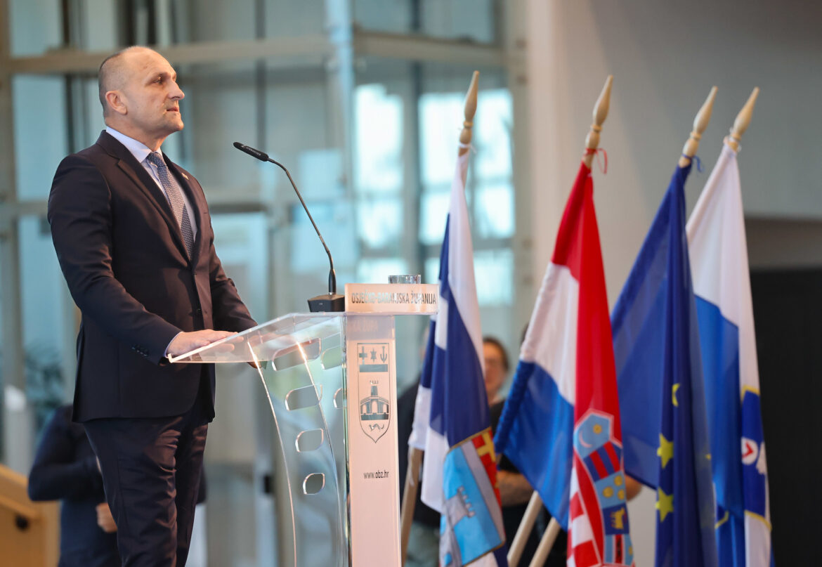 Man in a dark suit speaking at a podium during a formal event, multiple flags in the background