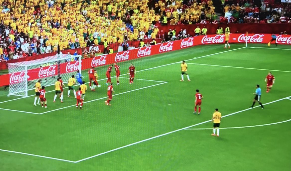 Players in yellow attack near the goal as red defenders and a referee watch on the green pitch. The stands are full of yellow-clad fans and Coca‑Cola ads line the sideline.