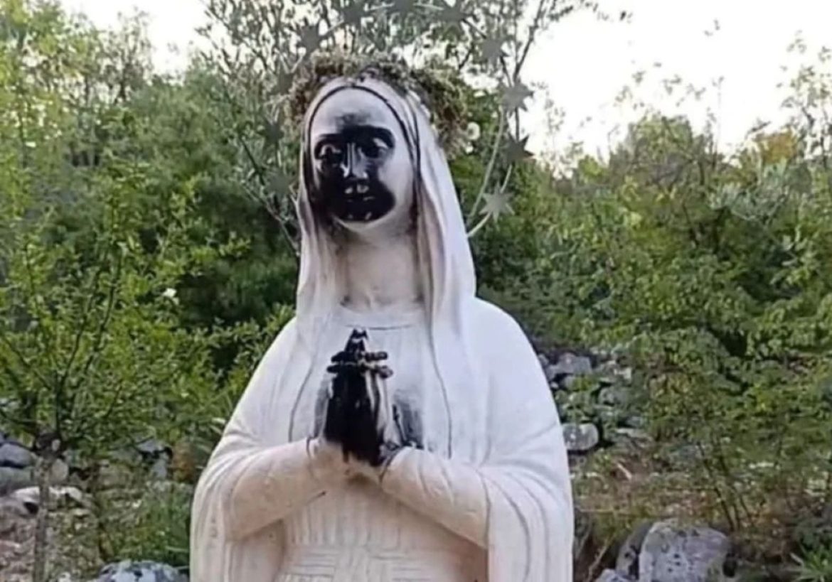Garden statue of a nun in a white robe, hands pressed in prayer, with a black-painted face amid greenery.