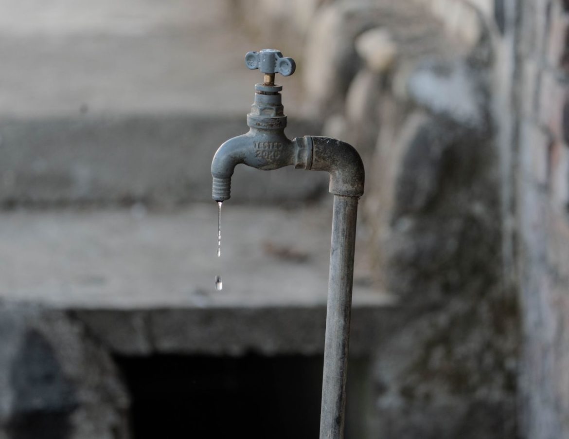 Rusty outdoor faucet dripping a thin stream of water against a blurred stone background.