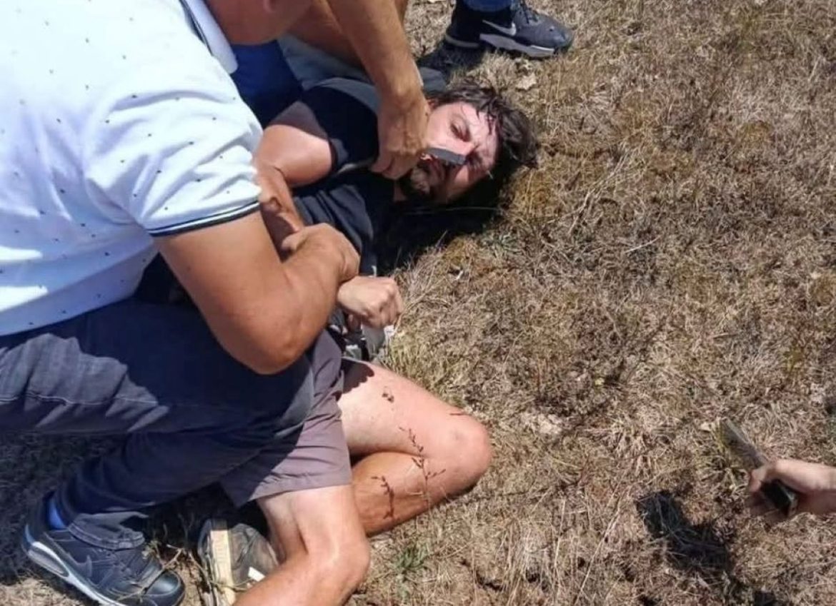 Man lying on dry grass while others hold him down, with visible leg injuries suggesting a rough outdoor incident.