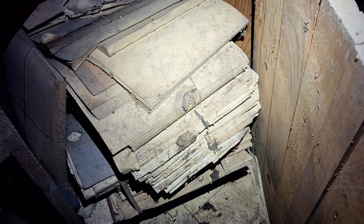 Pile of dusty, stacked slate tiles leaning against a wooden wall in a dim space, likely old roofing material.