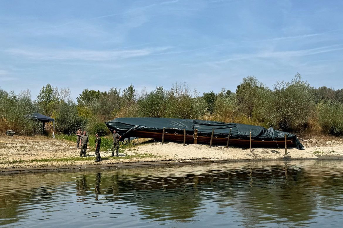 Group of soldiers standing on a sandy riverbank next to a large, tarp-covered boat.