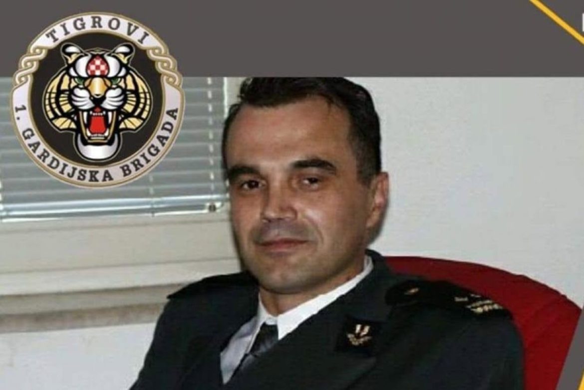 Man in a formal military uniform seated in an office, with a tiger-brigade insignia in the upper-left corner