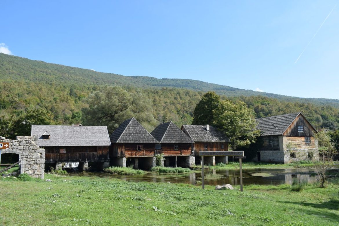 Row of wooden stilt houses by a calm water body, with forested hills in the background.