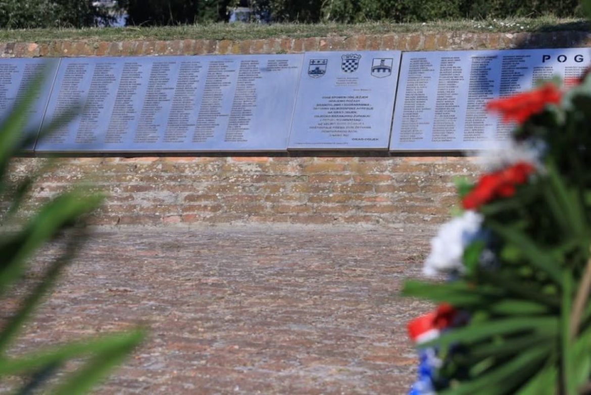 Outdoor memorial wall with metal plaques listing names, set on a brick base; a wreath of red, white, and blue flowers in the foreground.