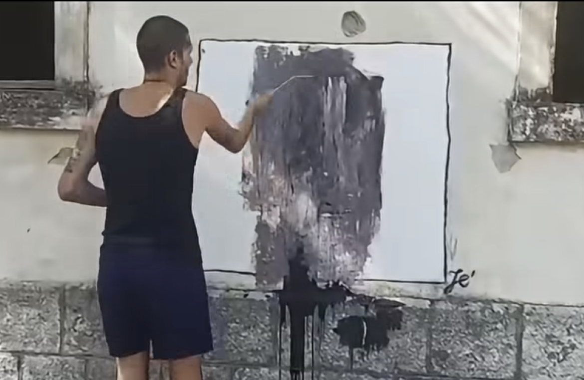 Man in a black tank top paints a dark abstract composition on a large white panel outdoors, with paint drips on the ground.