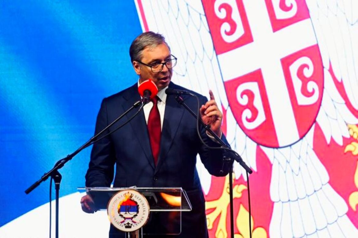 Man in a dark suit and red tie delivering a speech at a clear podium with a red microphone, in front of a large national crest backdrop.