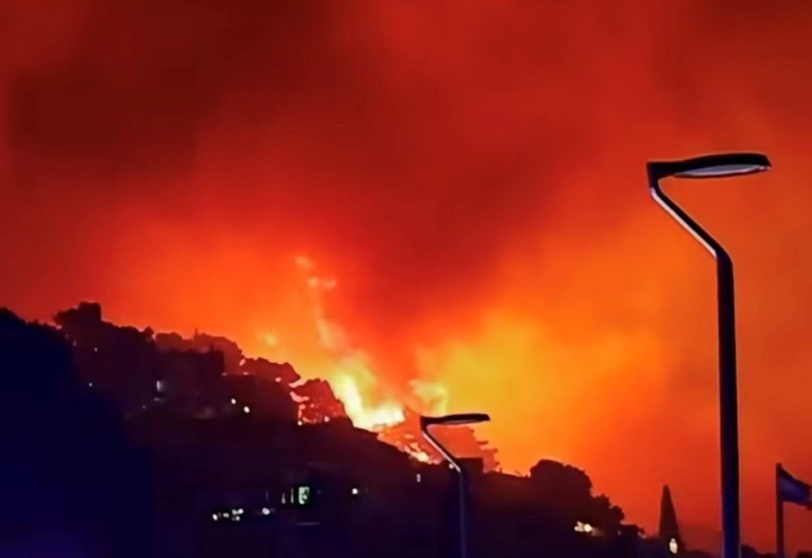 Nighttime wildfire lights up the hillside with large flames and dark smoke, streetlights visible in the foreground.